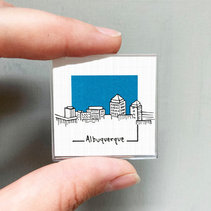 A person's hand holding a small square magnet with a drawing of a city skyline and the word "AlBUQUERQUE" written below it.