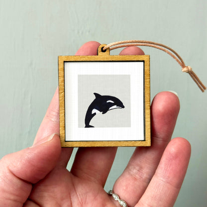 A person's hand holding a small wooden frame containing a black silhouette of a whale.