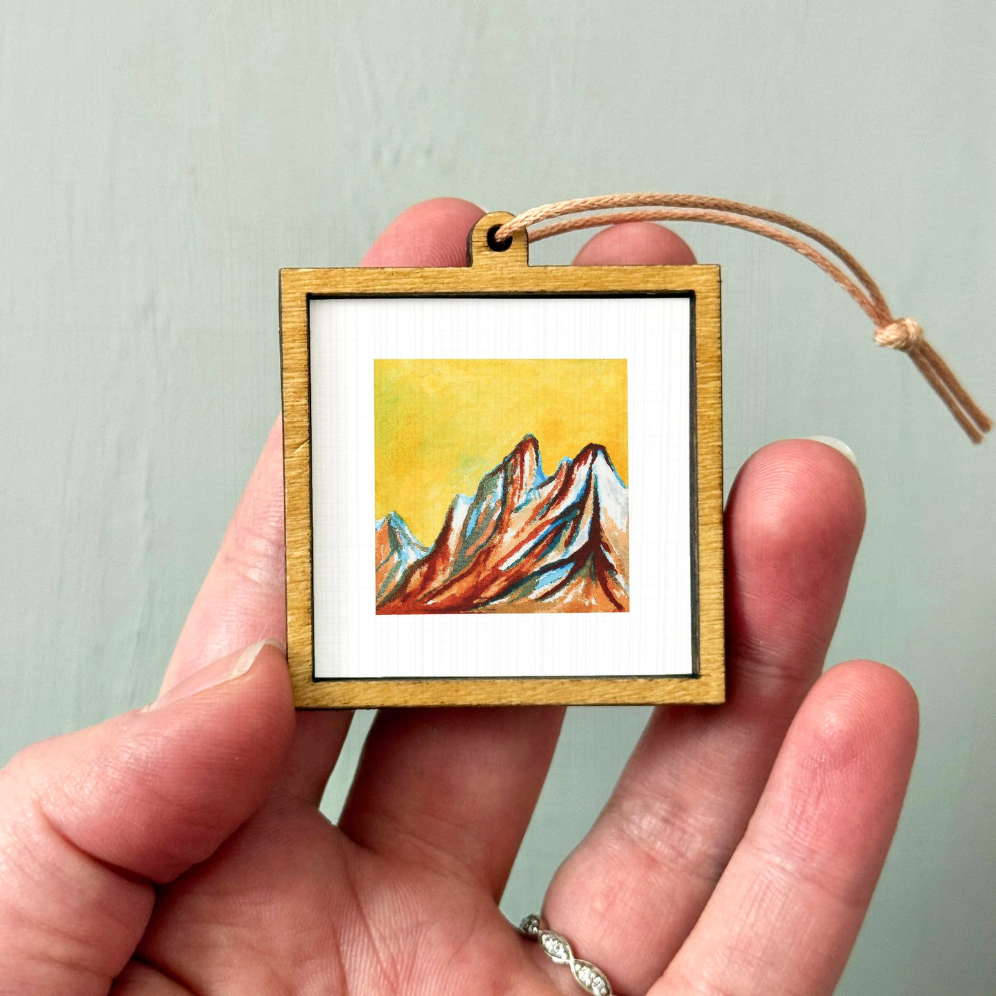 A hand holding a small wooden frame containing a painting of mountains.