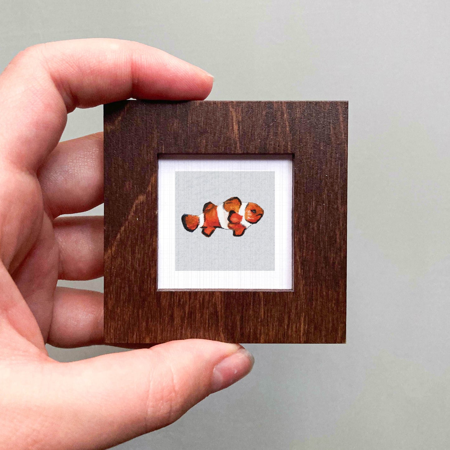 A hand is holding a small wooden frame containing a framed image of a clownfish.