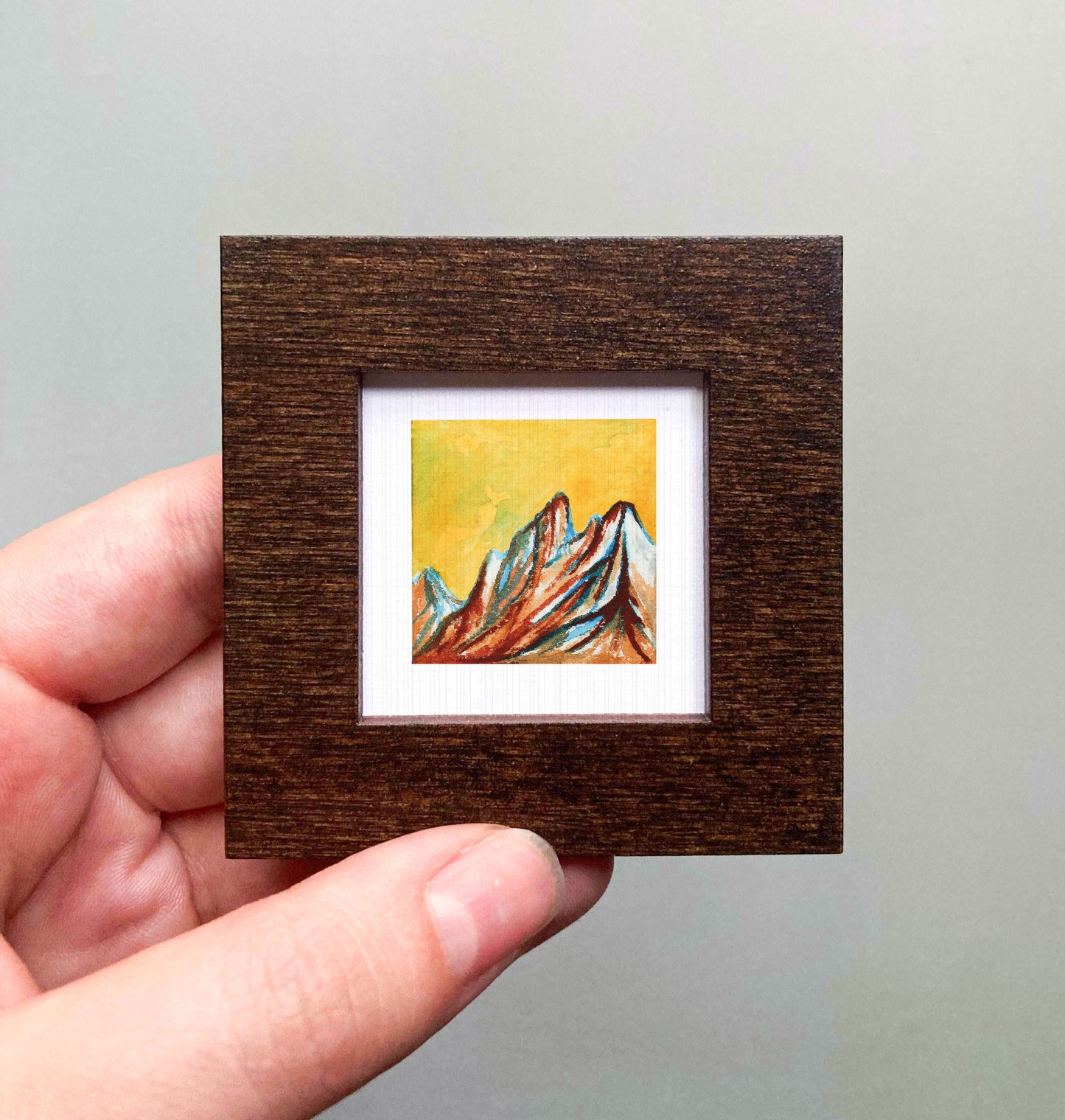 A hand is holding a small, square-shaped wooden frame containing a colorful painting of mountains.
