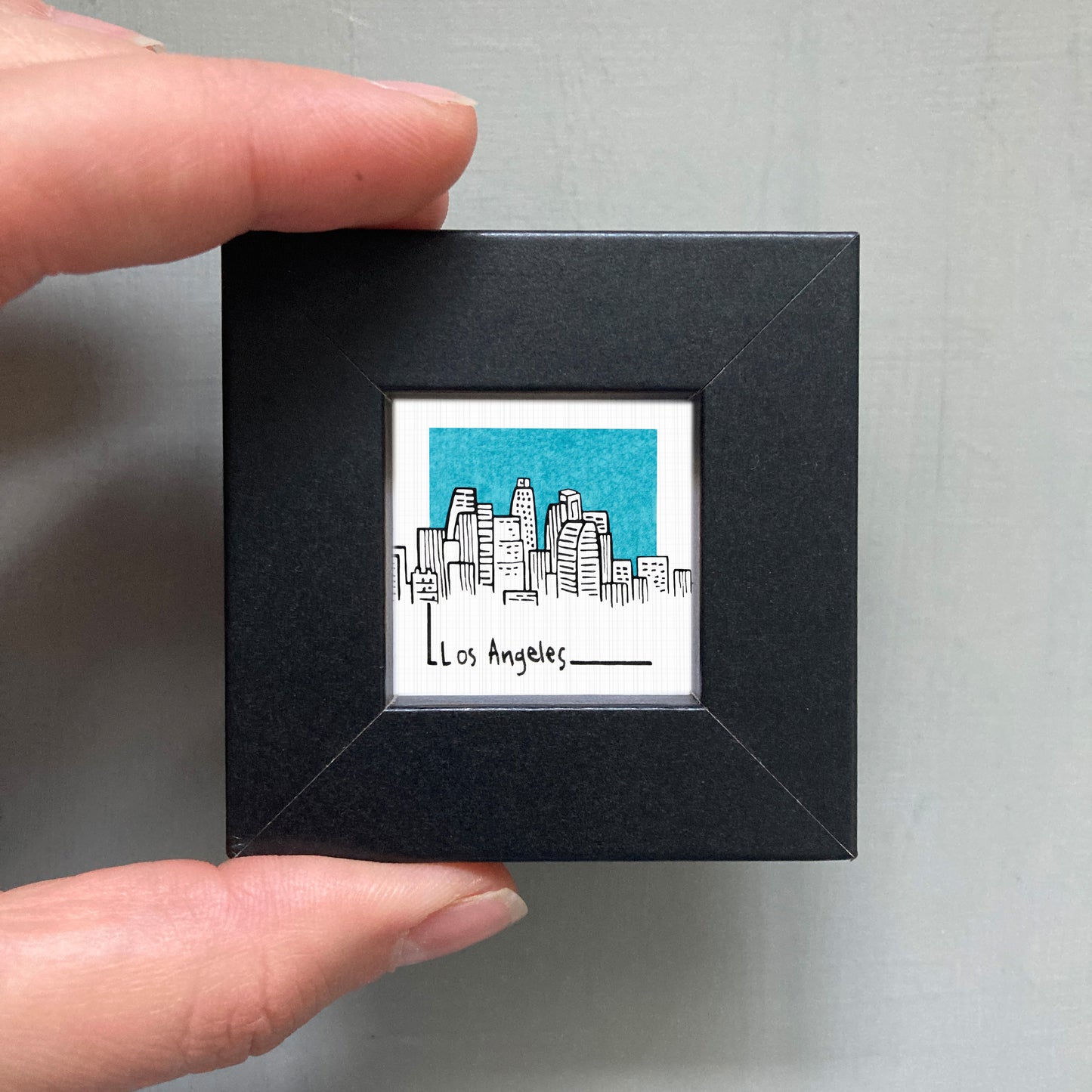 A person's hand holding a small black frame containing a drawing of a city skyline.
