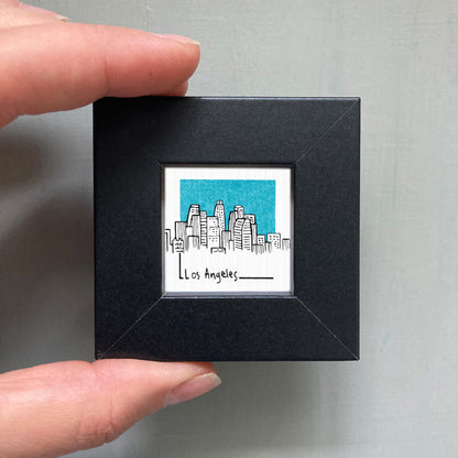 A person's hand holding a small black frame containing a drawing of a city skyline.