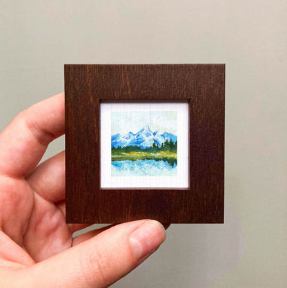 A hand is holding a small wooden frame containing a framed landscape painting depicting a mountain scene with a lake and trees.
