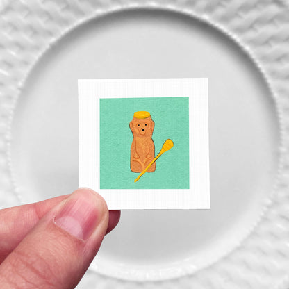 A hand is holding a small square with a picture of a brown bear wearing a yellow hat and holding a yellow object, which appears to be a toothbrush.