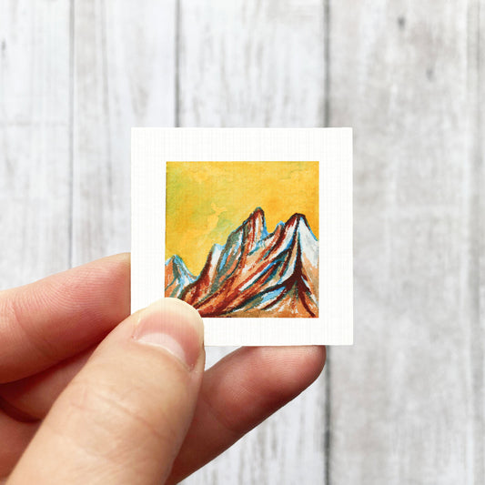A hand is holding a small square artwork depicting a mountain landscape with vibrant colors.