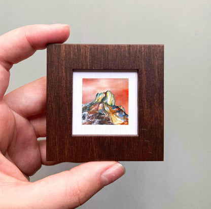 A hand is holding a small wooden frame containing a watercolor painting of a mountain landscape.