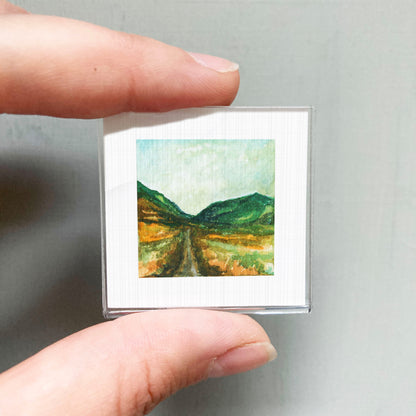 A person's hand holding a small square picture of a landscape with mountains and a winding road.