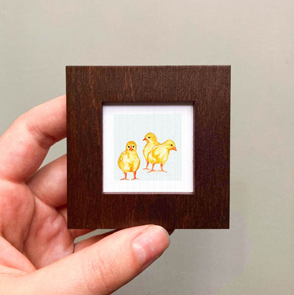 A hand is holding a small wooden frame containing a painting of three yellow chicks.