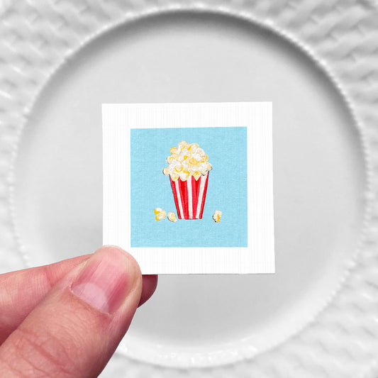 A hand is holding a small square with a picture of a red and white striped popcorn bucket on a blue background.