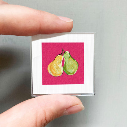 A person's hand holding a small square with a vibrant illustration of two pears on a pink background.