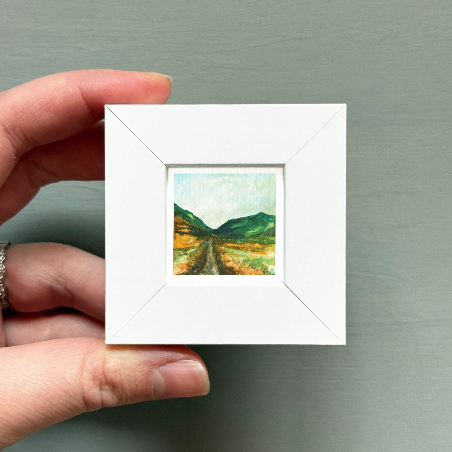 A hand is holding a small white square frame containing a painting of a landscape with mountains and a winding road.