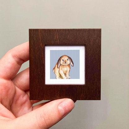 A hand is holding a small wooden frame containing a watercolor painting of a rabbit.