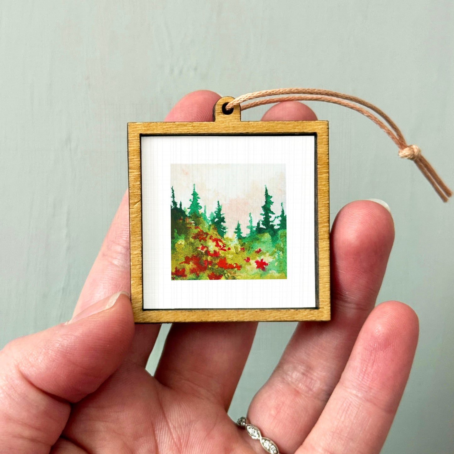 A hand holding a small wooden frame containing a painting of a forest scene with red flowers.