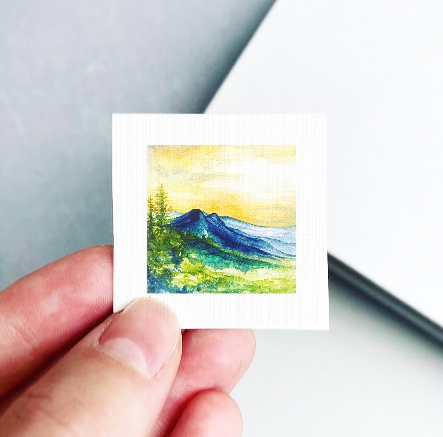 A hand is holding a small square piece of paper with a watercolor painting of a mountain landscape on it.