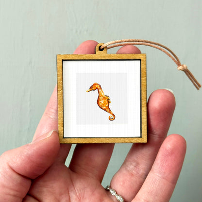 A person's hand holding a small wooden frame containing a picture of a seahorse.