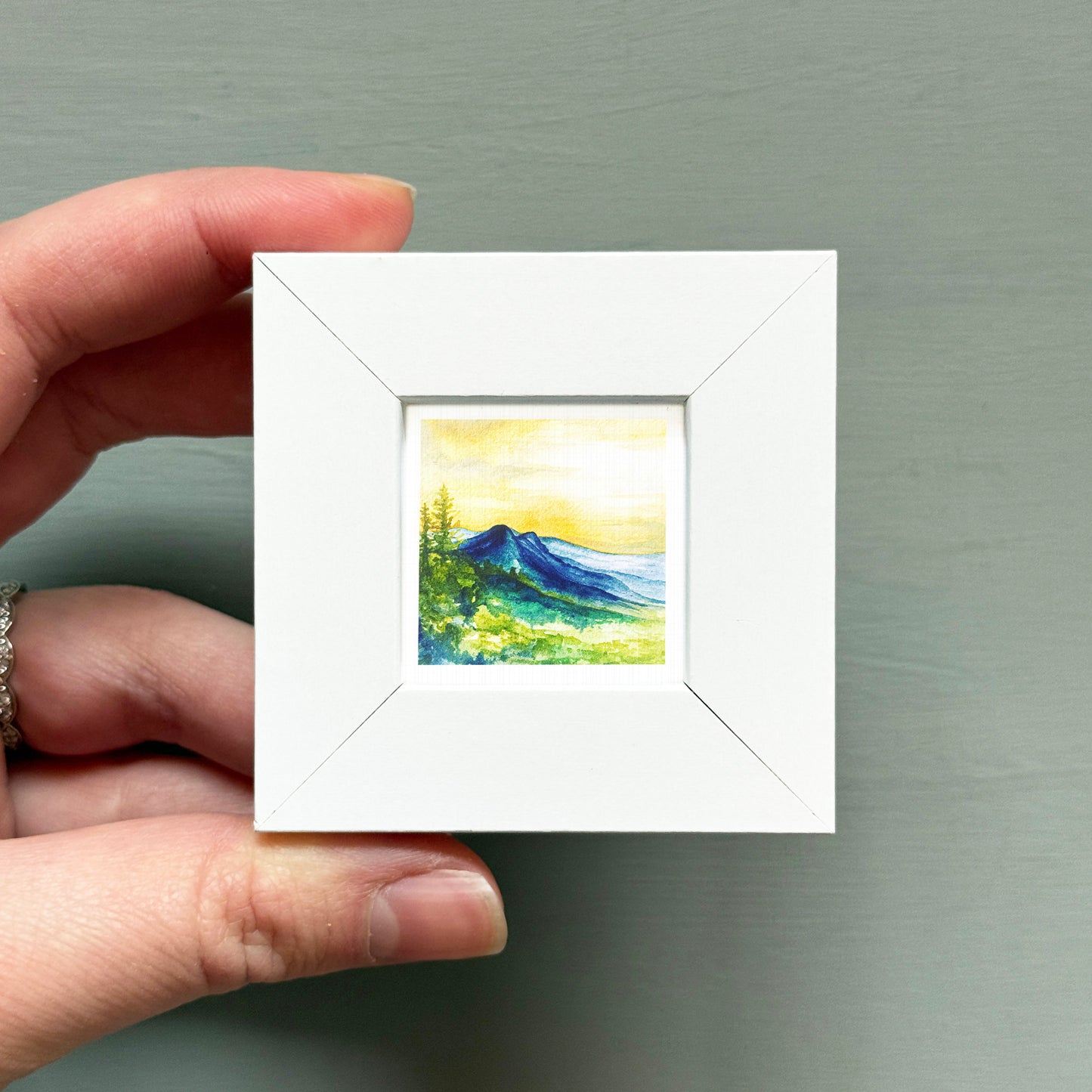 A hand is holding a small white square frame containing a watercolor painting of a mountain landscape.
