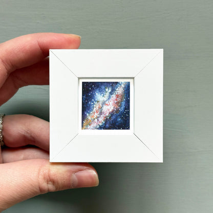 A hand is holding a small white square frame containing a painting of a galaxy.