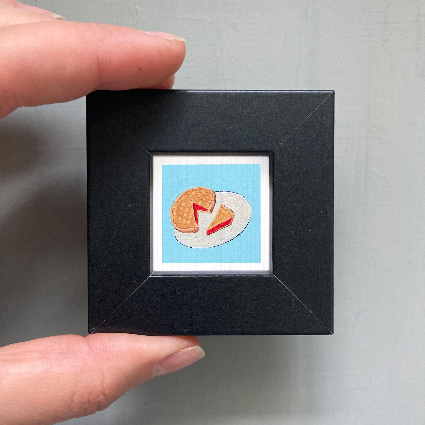 A hand is holding a small black frame containing a framed illustration of a piece of food, likely a slice of pie or waffle.