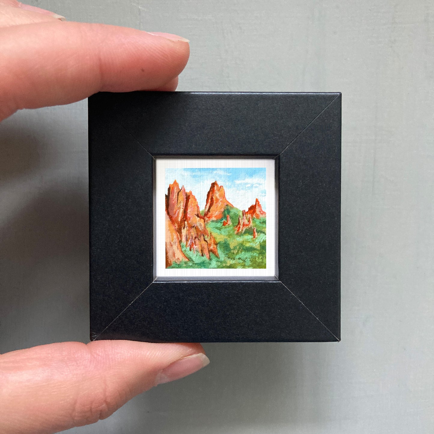 A person's hand holding a small black frame containing a framed painting of a mountain landscape.