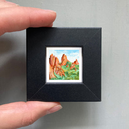 A person's hand holding a small black frame containing a framed painting of a mountain landscape.