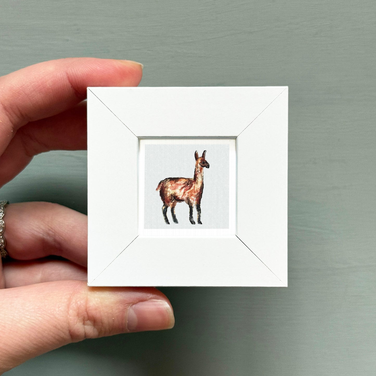 A hand is holding a small white square frame containing a detailed embroidered image of a llama.