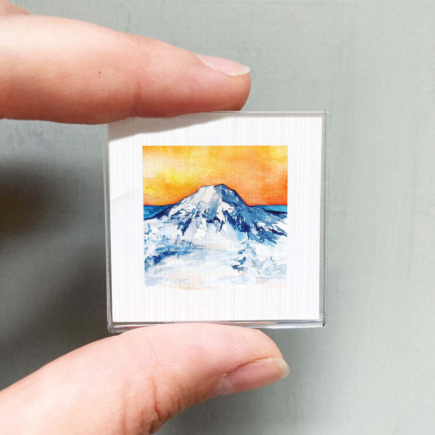 A person's hand is holding a small square picture of a mountain landscape with a vibrant orange and blue sky.