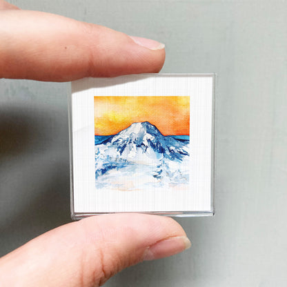 A person's hand is holding a small square picture of a mountain landscape with a vibrant orange and blue sky.