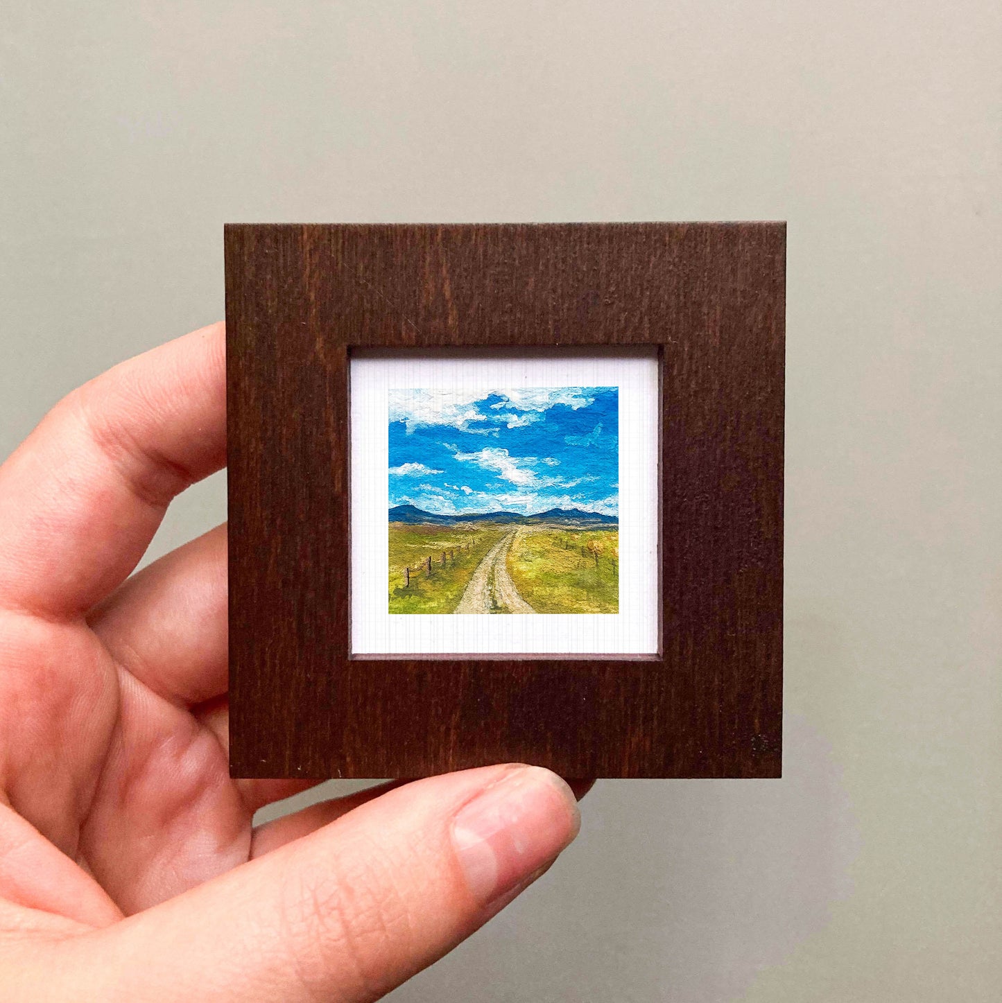 A hand is holding a small wooden frame containing a miniature landscape painting depicting a winding road leading to a blue sky with clouds.