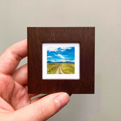 A hand is holding a small wooden frame containing a miniature landscape painting depicting a winding road leading to a blue sky with clouds.