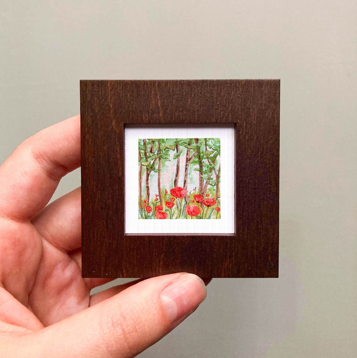 A hand is holding a small, square-shaped wooden frame containing a painting of red flowers in a forest.