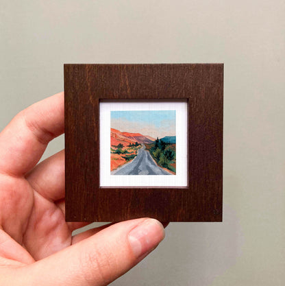 A hand is holding a small wooden frame containing a painting of a winding road through a desert landscape.