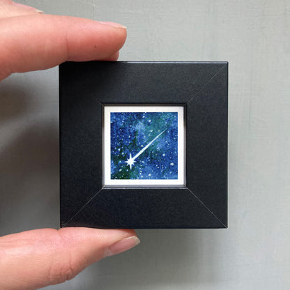 A hand is holding a small black frame containing a framed image of a starry night sky.
