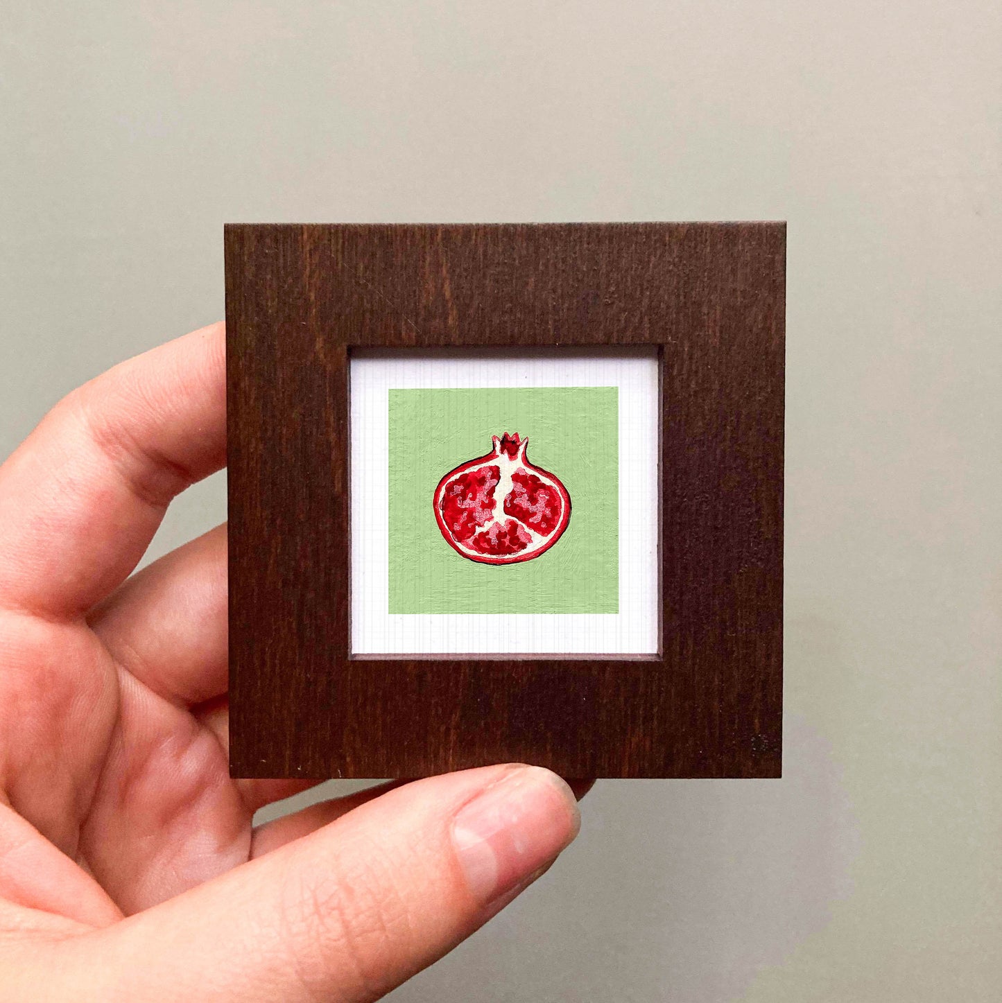 A hand is holding a small wooden frame containing a framed image of a pomegranate fruit.
