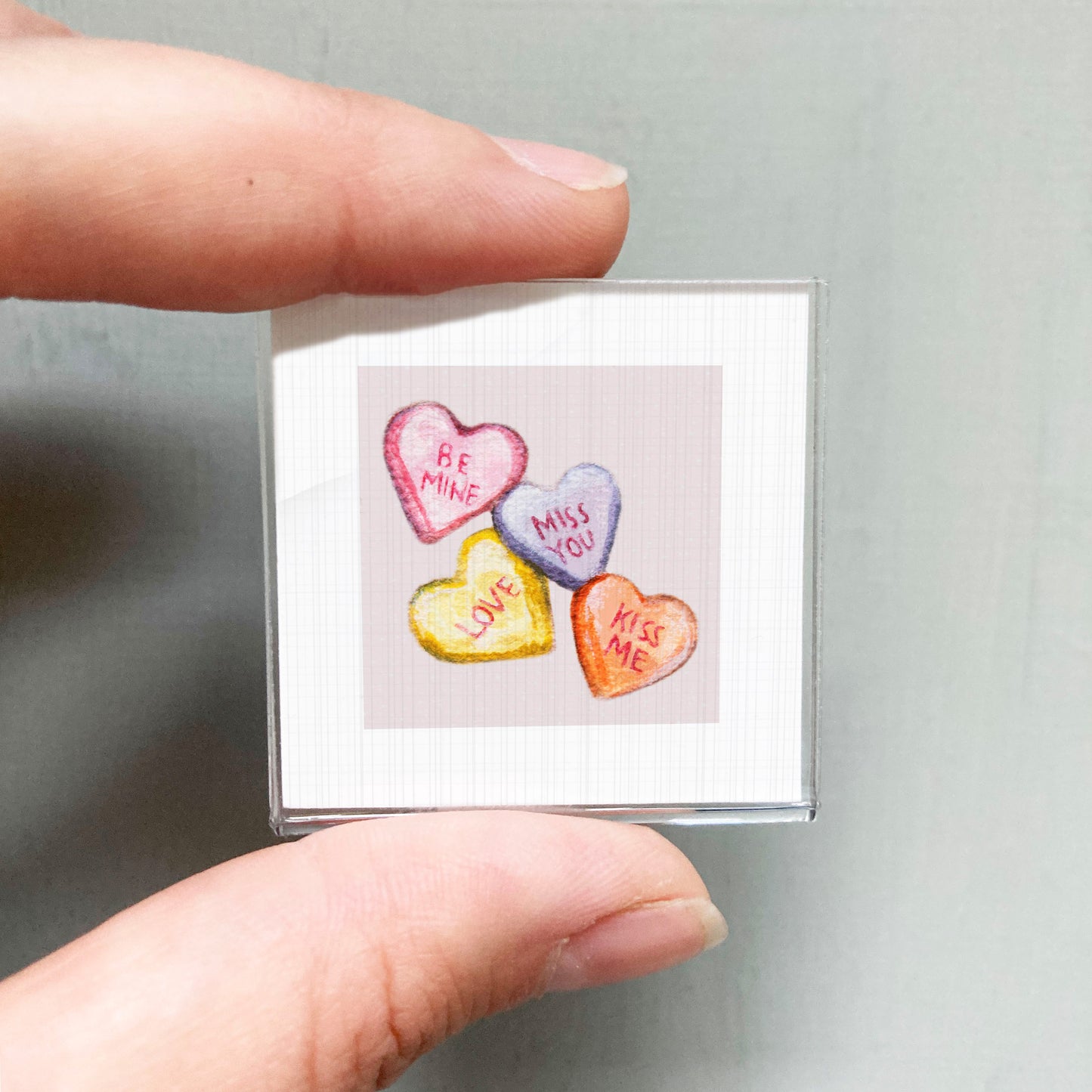 A person's hand holding a small square picture with four heart-shaped candies on it, each with a different message written on it.
