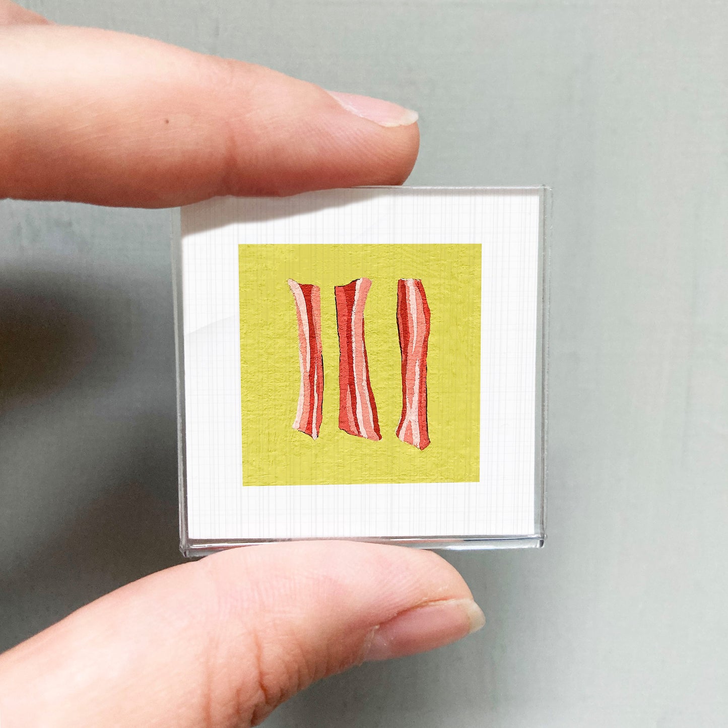 A person's hand holding a small square with a yellow background and three red lines, resembling a graphic design or illustration.