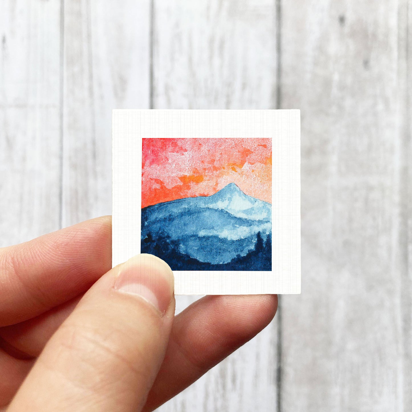 A hand is holding a small square artwork depicting a mountain landscape with a vibrant sunset sky.