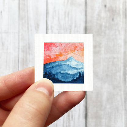 A hand is holding a small square artwork depicting a mountain landscape with a vibrant sunset sky.