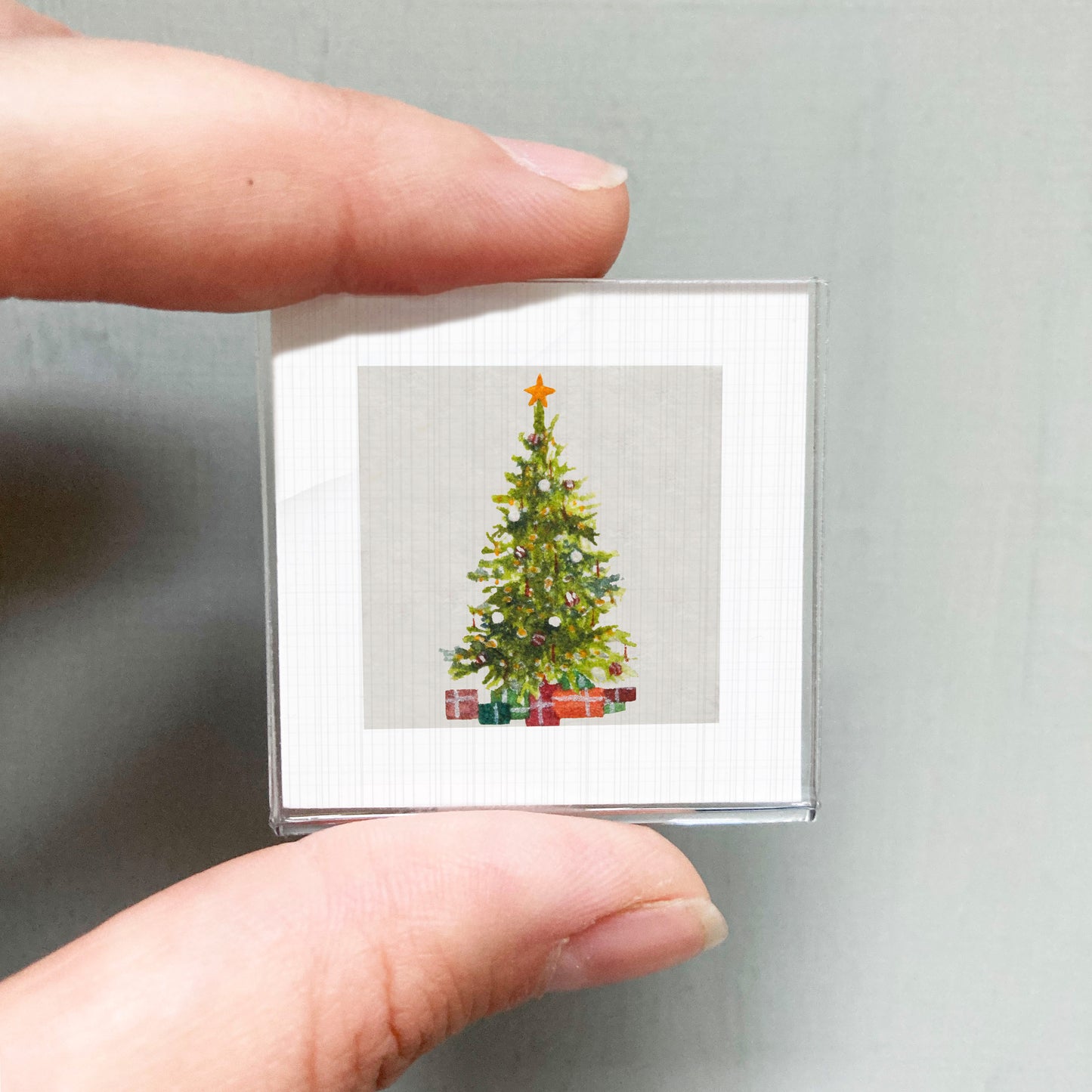 A hand holding a small square picture of a Christmas tree with presents underneath.
