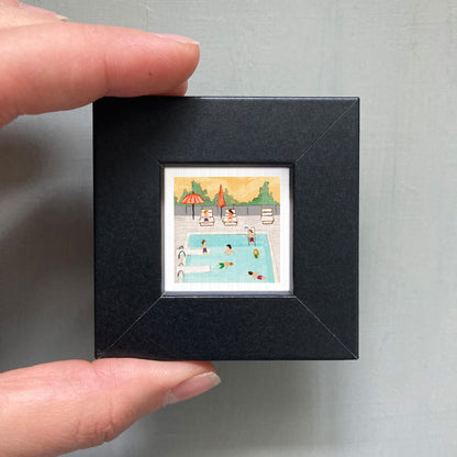 A person's hand is holding a small framed picture of a swimming pool scene with people enjoying the water and lounging on chairs.
