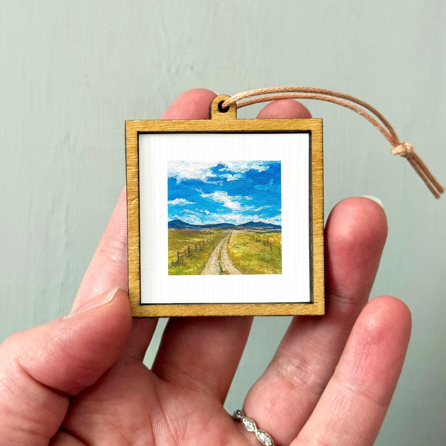 A person's hand holding a small wooden frame containing a framed landscape painting depicting a rural road leading to a blue sky with clouds.