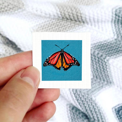A hand is holding a small square card with a colorful illustration of a butterfly on it.