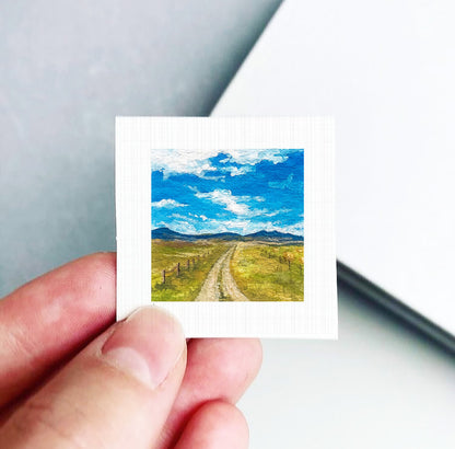 A hand holding a small square picture depicting a rural landscape with a dirt road leading through a grassy field and mountains in the distance.