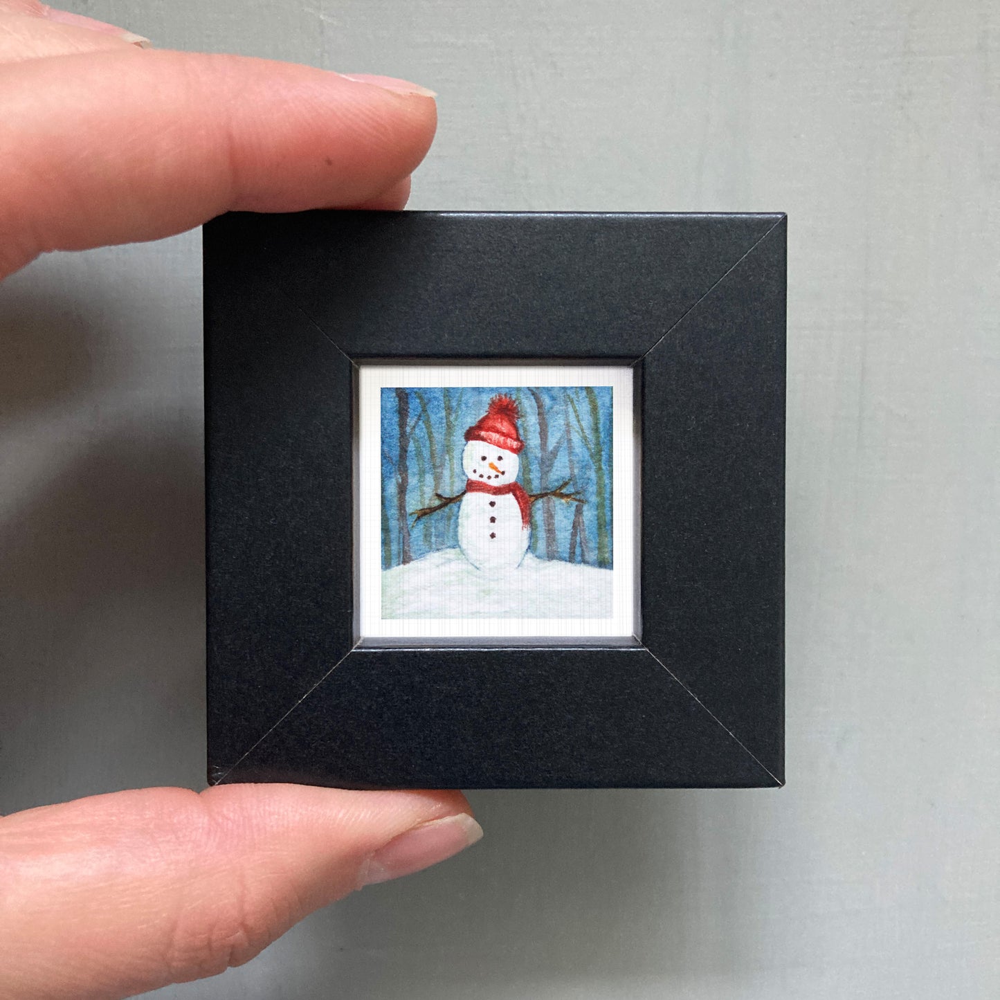 A person's hand is holding a small black picture frame containing a miniature painting of a snowman.