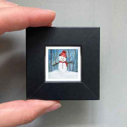 A person's hand is holding a small black picture frame containing a miniature painting of a snowman.