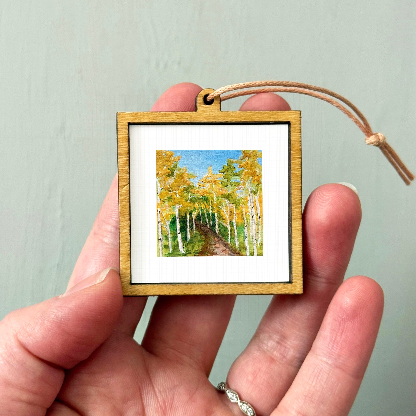 A hand holding a small wooden frame containing a painting of a forest with tall trees and a winding path.