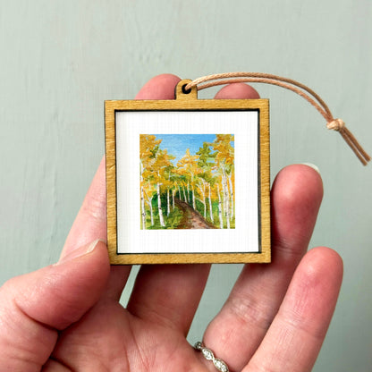 A hand holding a small wooden frame containing a painting of a forest with tall trees and a winding path.