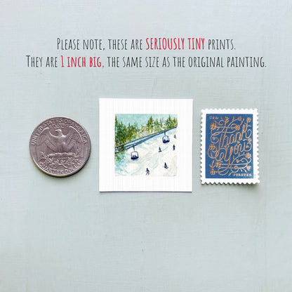 three small prints: a silver coin, a white square print of a snowy landscape with ski lifts, and a blue postcard with gold lettering.