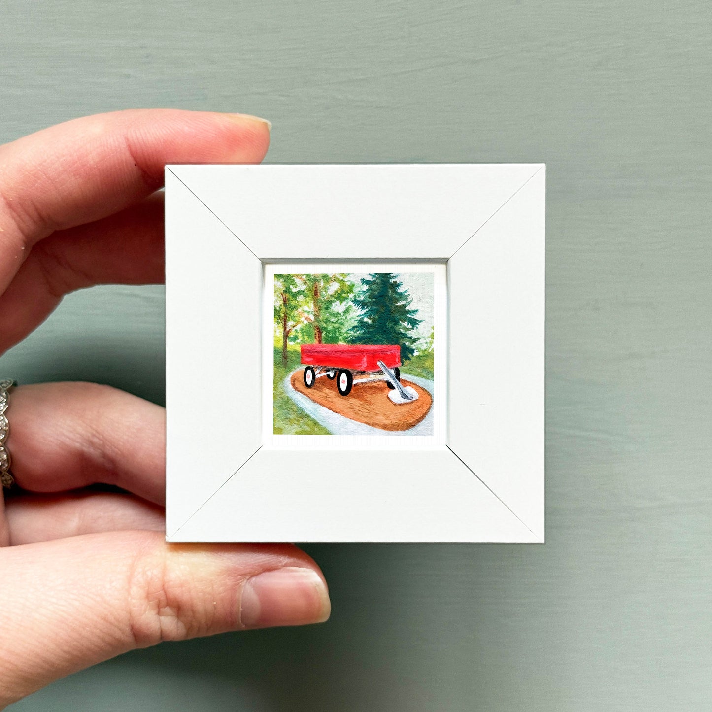 A hand is holding a small white picture frame containing a painting of a red wagon on a dirt path surrounded by trees.