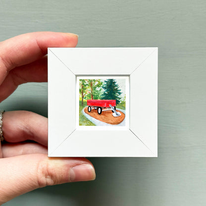 A hand is holding a small white picture frame containing a painting of a red wagon on a dirt path surrounded by trees.